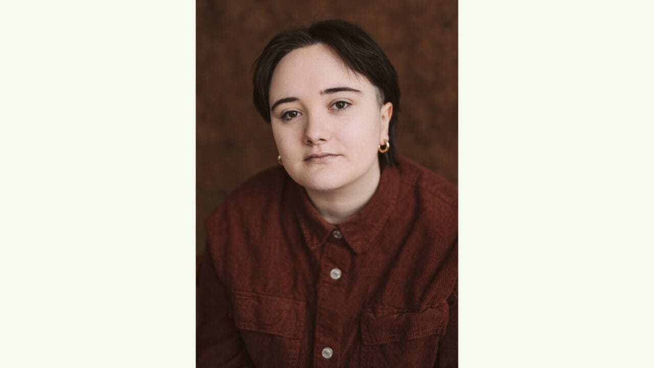A person with short dark hair, wearing a red button-up shirt and small hoop earrings, looks directly at the camera against a brown background.