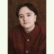 A person with short dark hair, wearing a red button-up shirt and small hoop earrings, looks directly at the camera against a brown background.