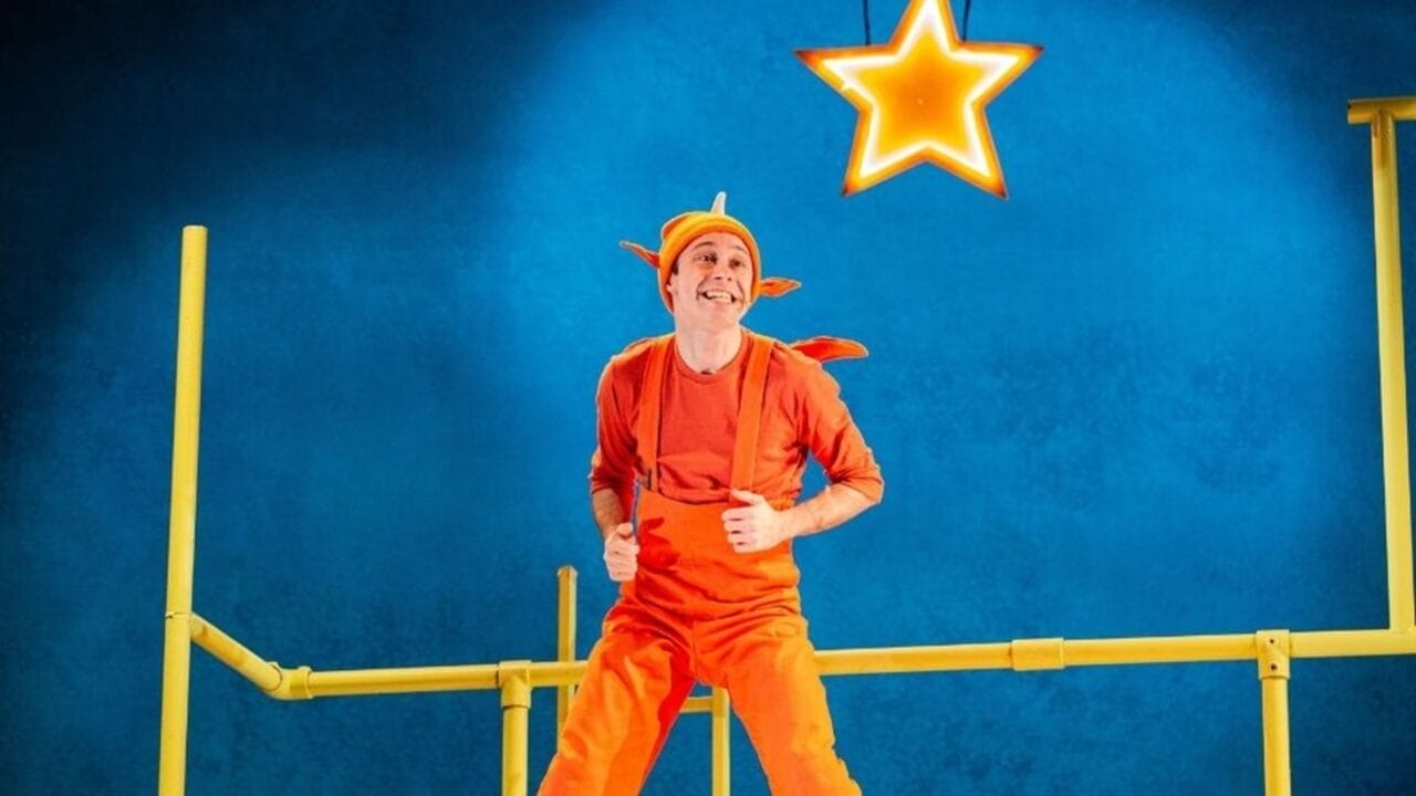 A person in an orange costume with a hat resembling an antenna stands on a yellow structure, smiling in the spotlight, with a glowing star suspended above them against a blue background.