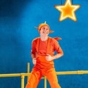 A person in an orange costume with a hat resembling an antenna stands on a yellow structure, smiling in the spotlight, with a glowing star suspended above them against a blue background.