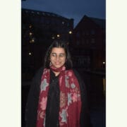 A person with glasses and a red floral scarf stands outside at night in front of buildings with lit windows.