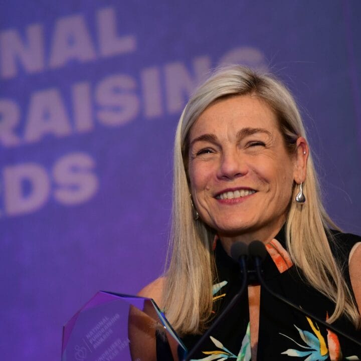 A woman smiles at a podium holding an award, with a "National Fundraising Awards" sign in the background.