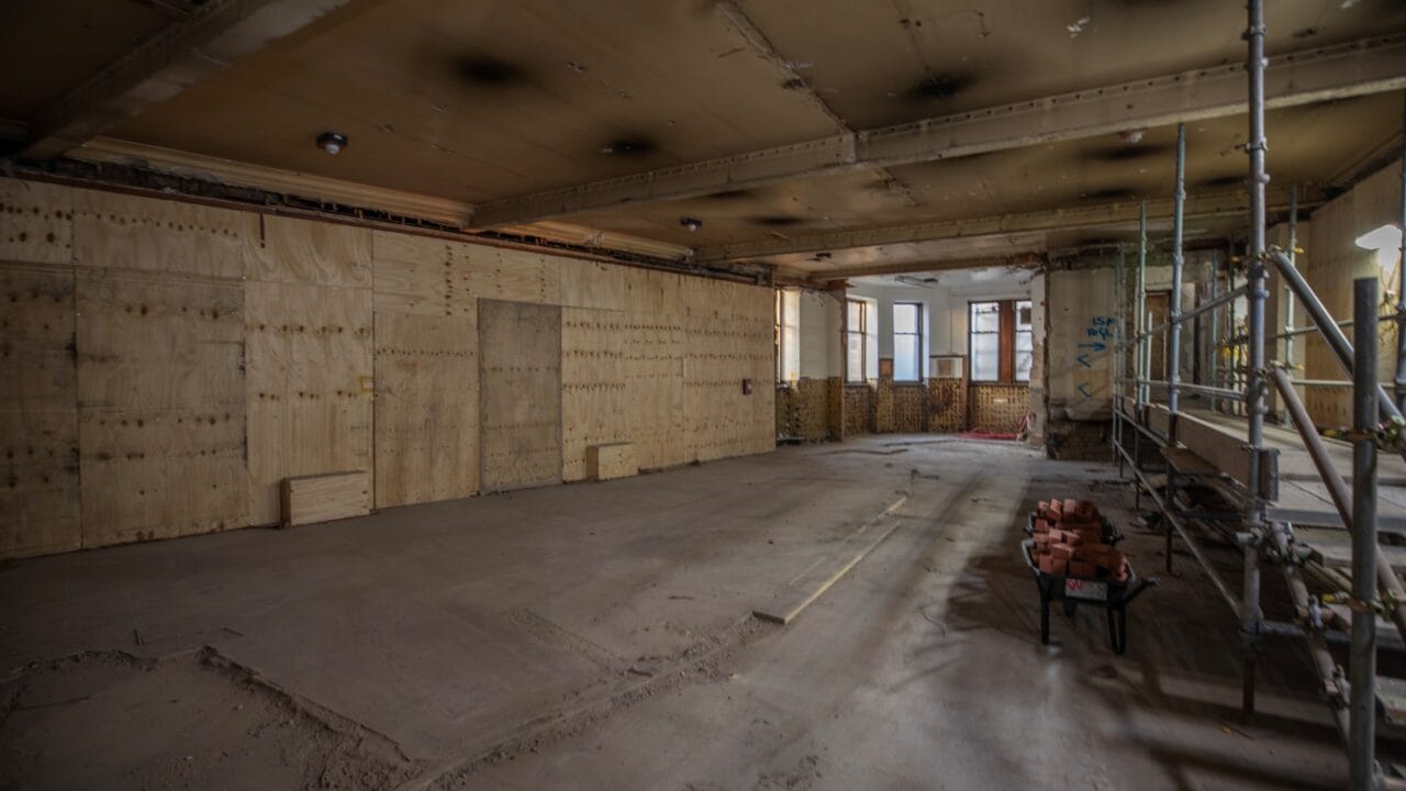 Large empty room under renovation with bare plywood walls, exposed ceiling, scattered construction materials, and scaffold on the right. Natural light enters through windows at the back.