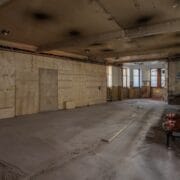 Large empty room under renovation with bare plywood walls, exposed ceiling, scattered construction materials, and scaffold on the right. Natural light enters through windows at the back.