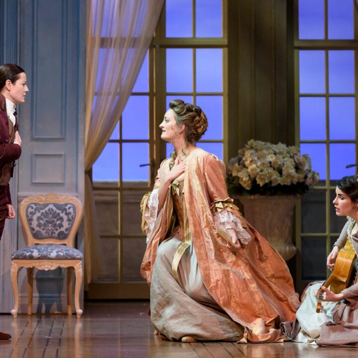 Three actors in period costumes perform on a stage set resembling an elegant 18th-century room; one stands, one sits on a couch, and one sits on the floor playing a guitar.