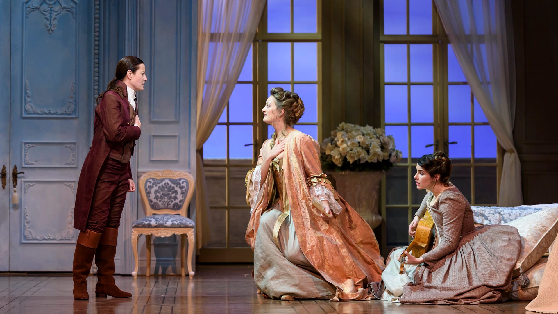 Three actors in period costumes perform on a stage set resembling an elegant 18th-century room; one stands, one sits on a couch, and one sits on the floor playing a guitar.