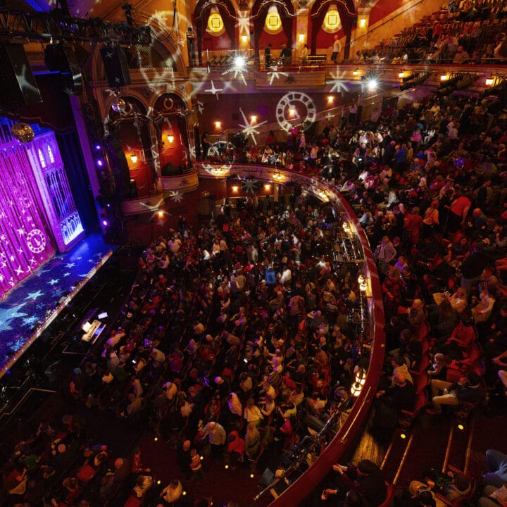 A crowded theater auditorium with audience members seated, facing a stage lit with purple lighting and projected star patterns.