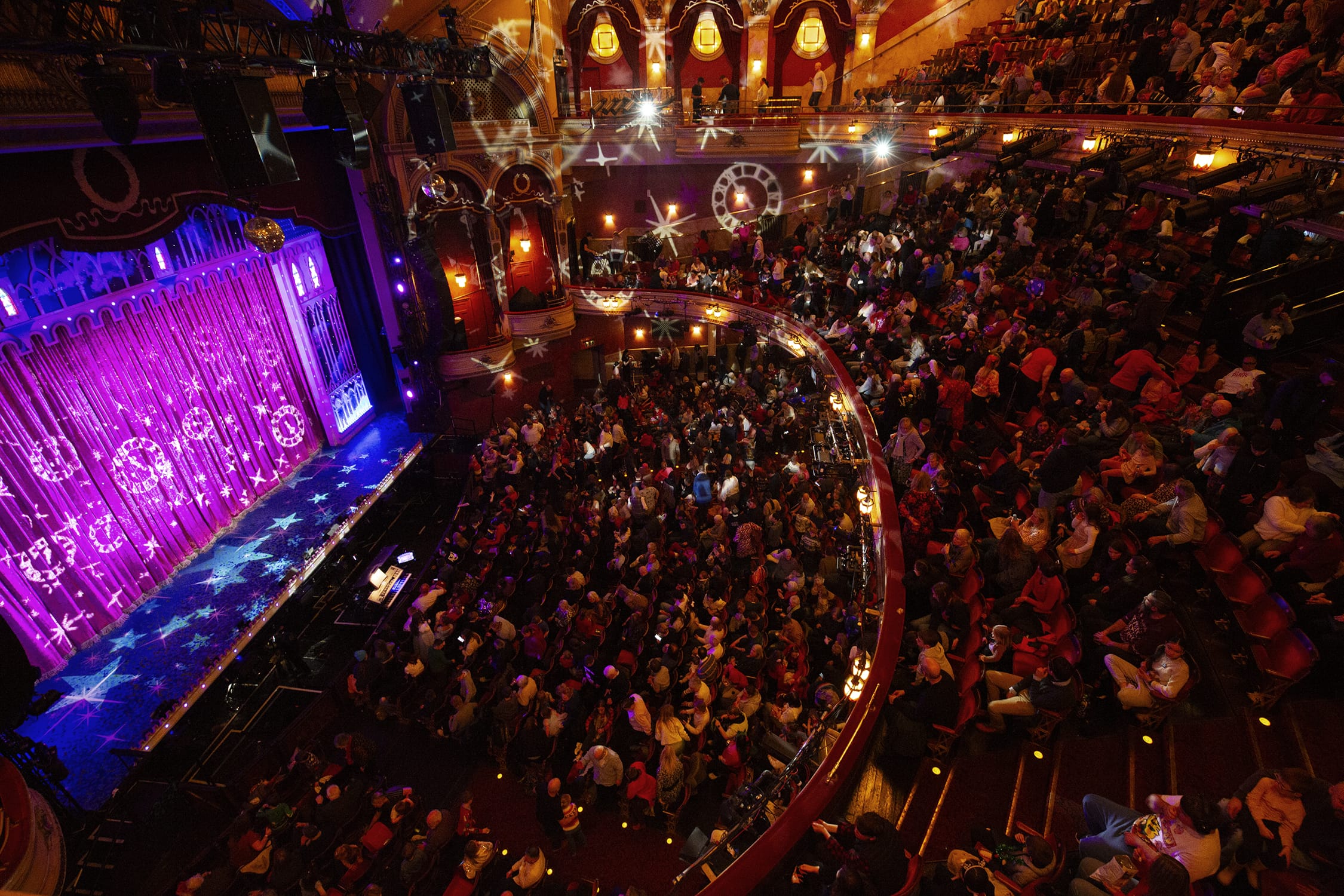 A crowded theater auditorium with audience members seated, facing a stage lit with purple lighting and projected star patterns.