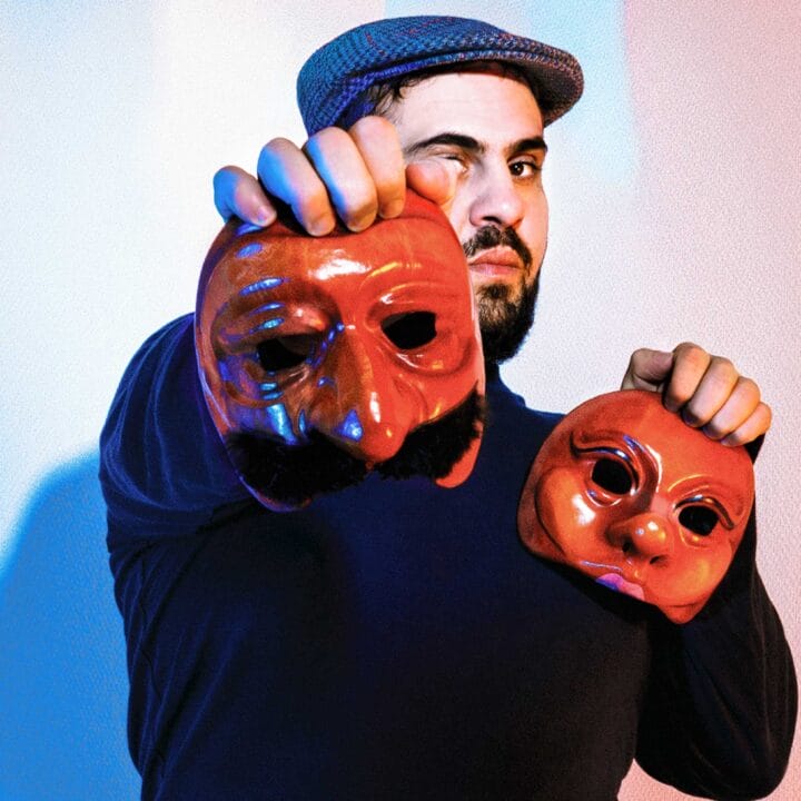 A man wearing a flat cap holds two theatrical masks, one in each hand, in front of a blue and pink lit background.