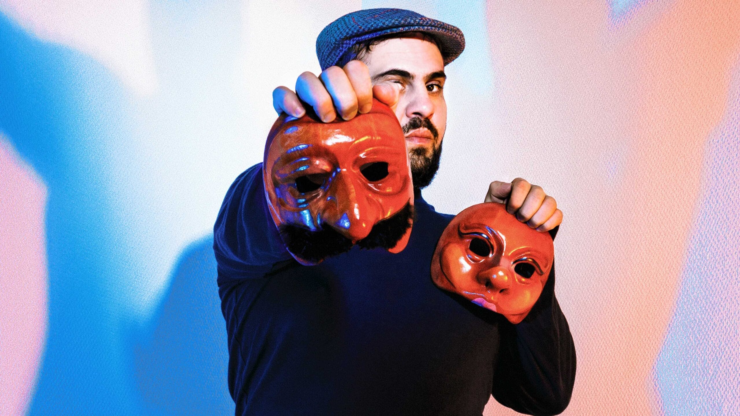 A man wearing a flat cap holds two theatrical masks, one in each hand, in front of a blue and pink lit background.