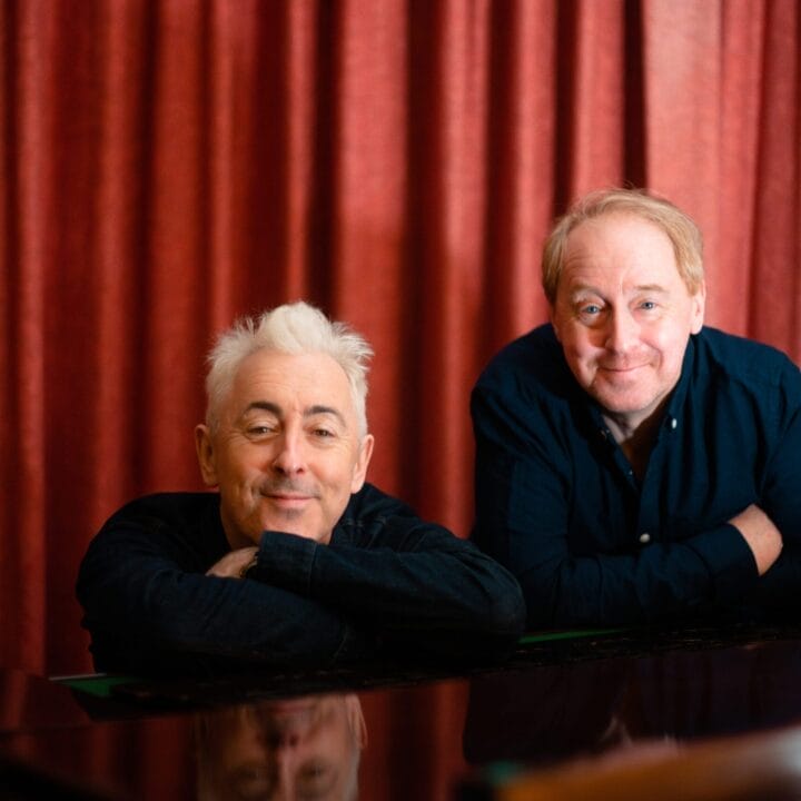 Two men with light skin rest their arms on a piano and smile at the camera. A red curtain hangs in the background, setting a stage-like scene reminiscent of a Festival Theatre during High Life moments.