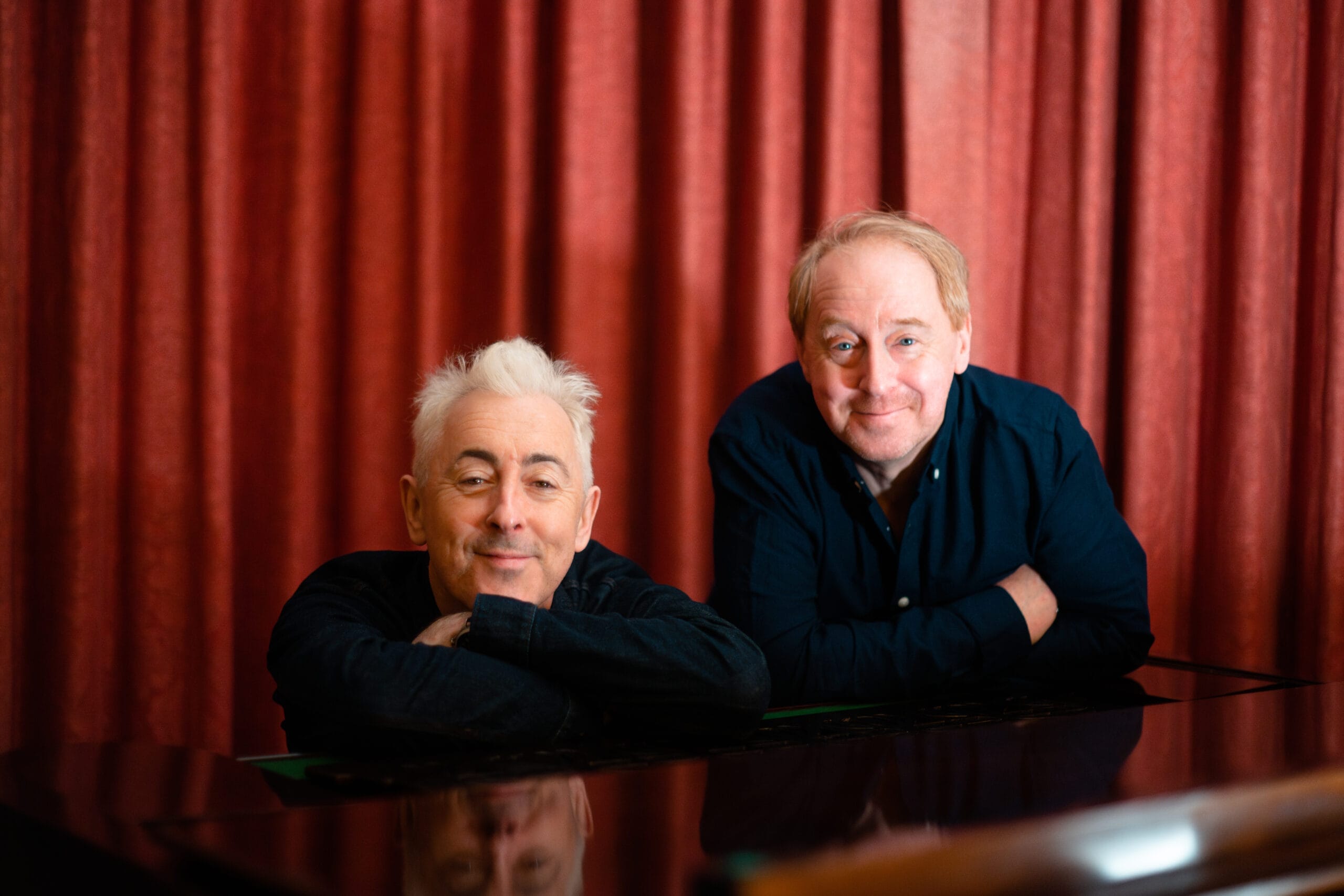 Two men with light skin rest their arms on a piano and smile at the camera. A red curtain hangs in the background, setting a stage-like scene reminiscent of a Festival Theatre during High Life moments.