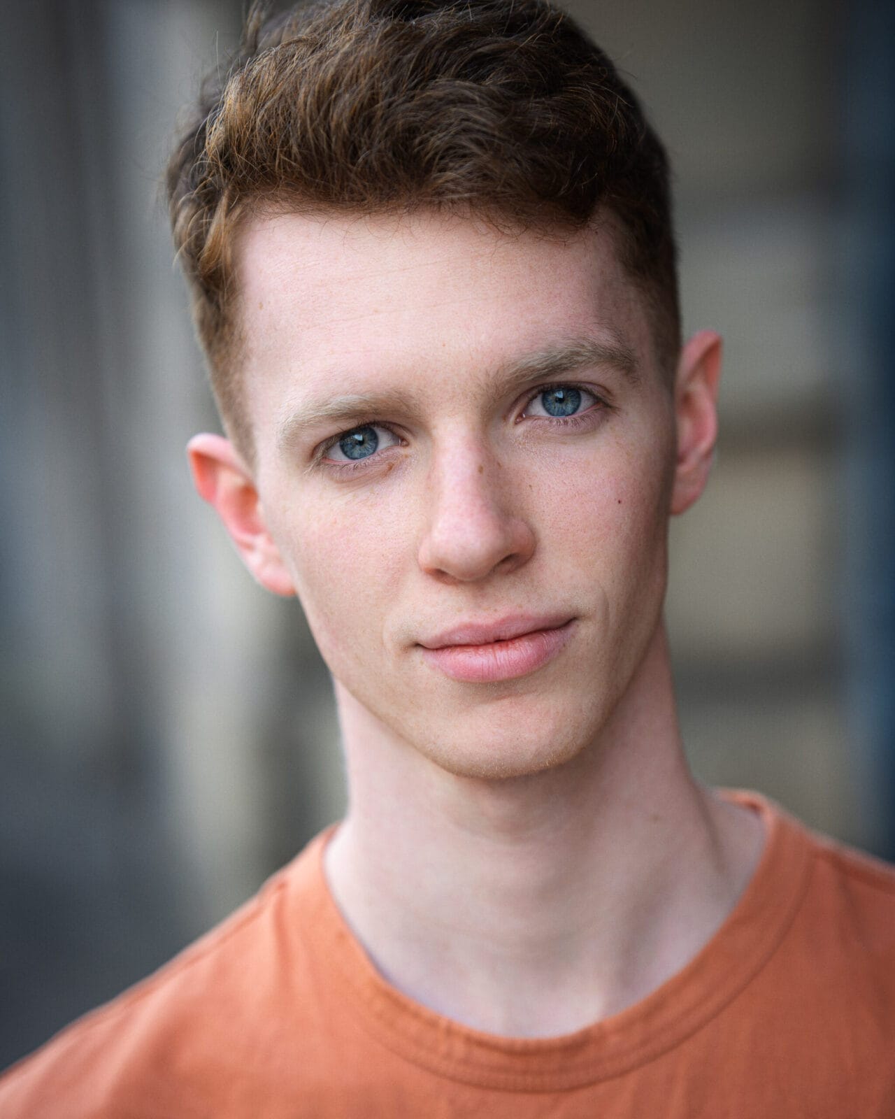 A young man with short brown hair and blue eyes wearing an orange shirt, looking directly at the camera with a neutral expression, as if ready to perform at Open Floor or take part in a Scratch Night event.