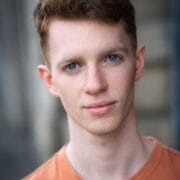 A young man with short brown hair and blue eyes wearing an orange shirt, looking directly at the camera with a neutral expression, as if ready to perform at Open Floor or take part in a Scratch Night event.