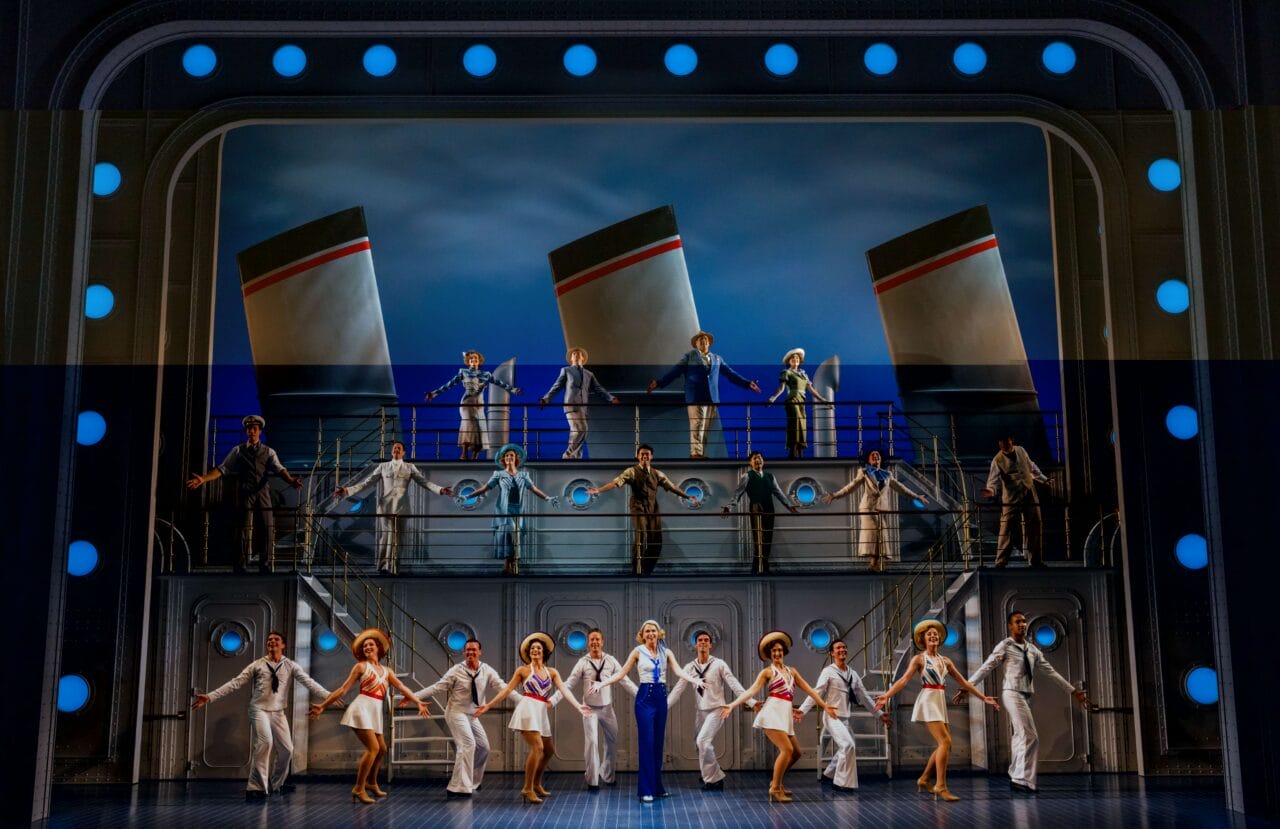 A large cast performs a musical number on a two-level stage set resembling a ship, with lifebuoys, sailor costumes, and smokestacks in the background.