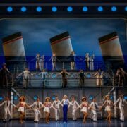 A large cast performs a musical number on a two-level stage set resembling a ship, with lifebuoys, sailor costumes, and smokestacks in the background.
