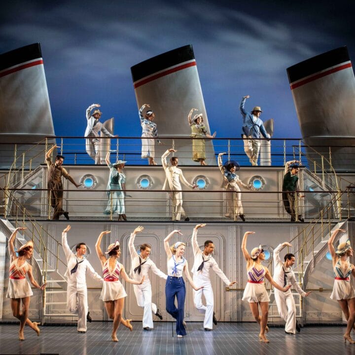 A large group of performers in nautical costumes dance and pose on a stage set resembling a cruise ship, with smokestacks and railings in the background.