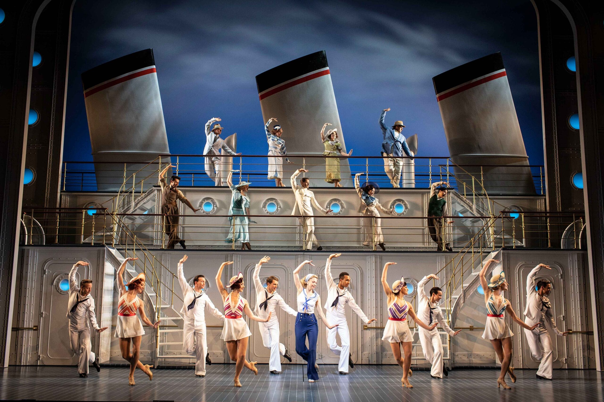 A large group of performers in nautical costumes dance and pose on a stage set resembling a cruise ship, with smokestacks and railings in the background.