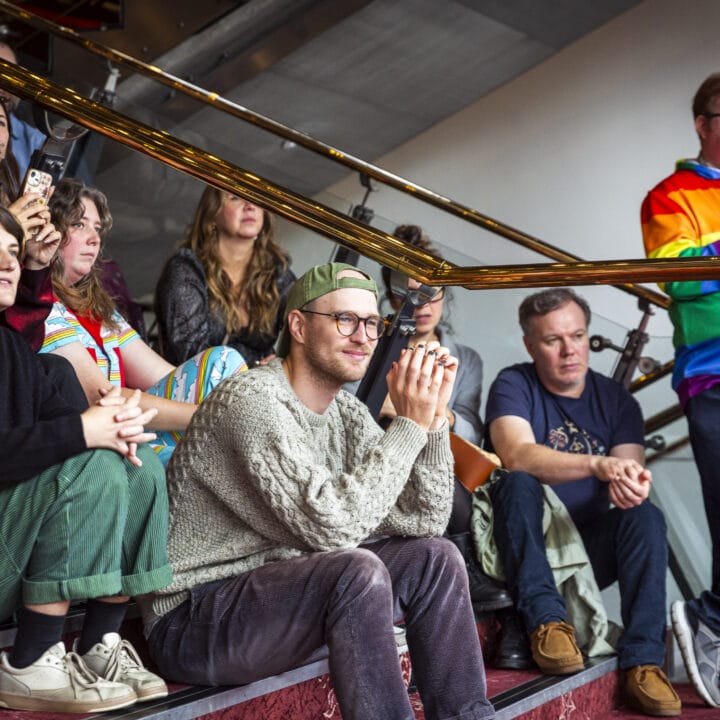A group of people sit and stand on stairs, attentively watching something off-camera. One person stands wearing a rainbow-colored jacket.