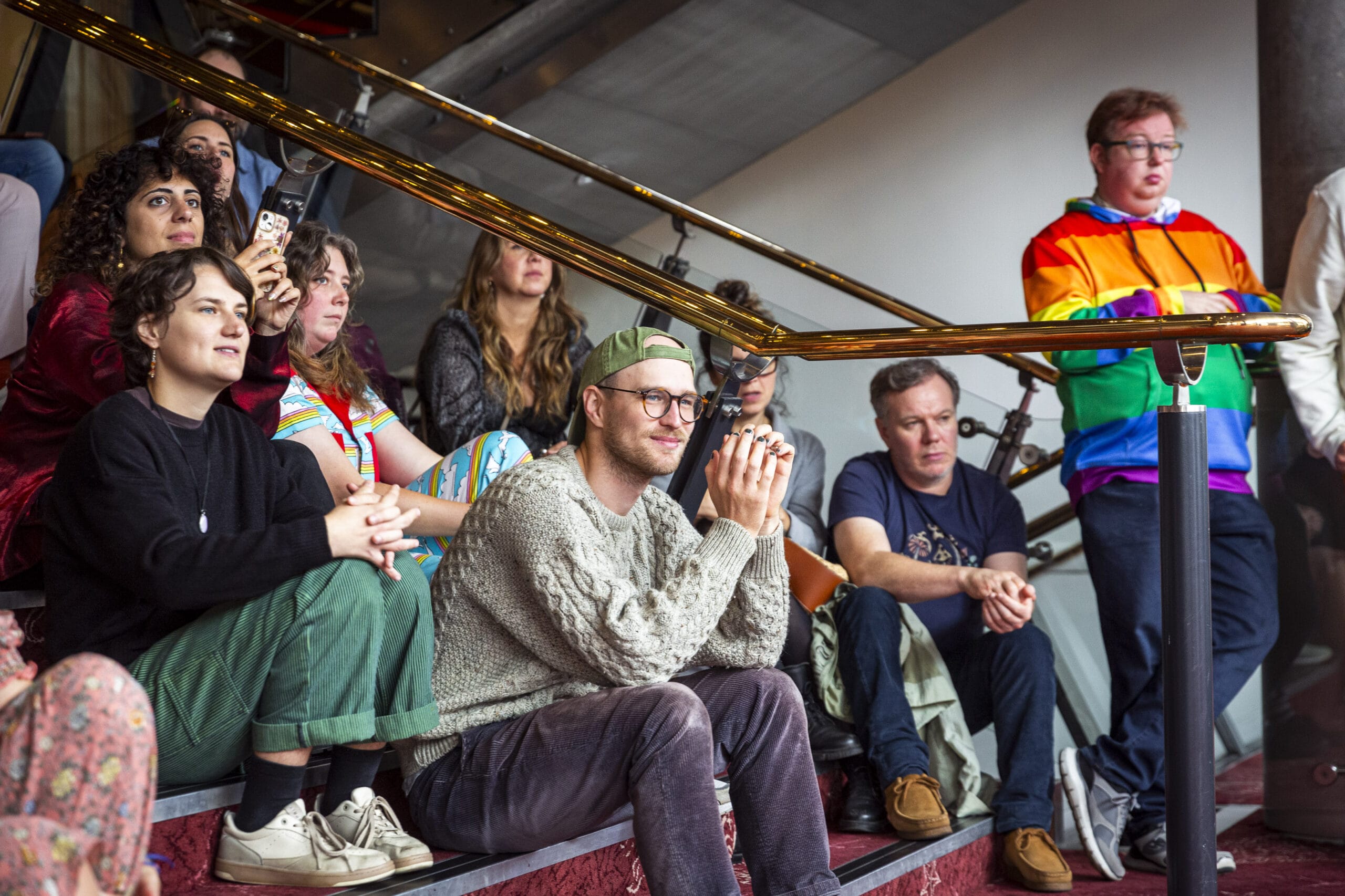 A group of people sit and stand on stairs, attentively watching something off-camera. One person stands wearing a rainbow-colored jacket.