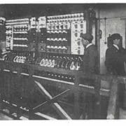 Black and white photo. Three people, including Bethia on the far left, stand in front of a large early 20th-century electrical control panel.