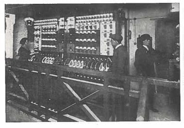 Black and white photo. Three people, including Bethia on the far left, stand in front of a large early 20th-century electrical control panel.