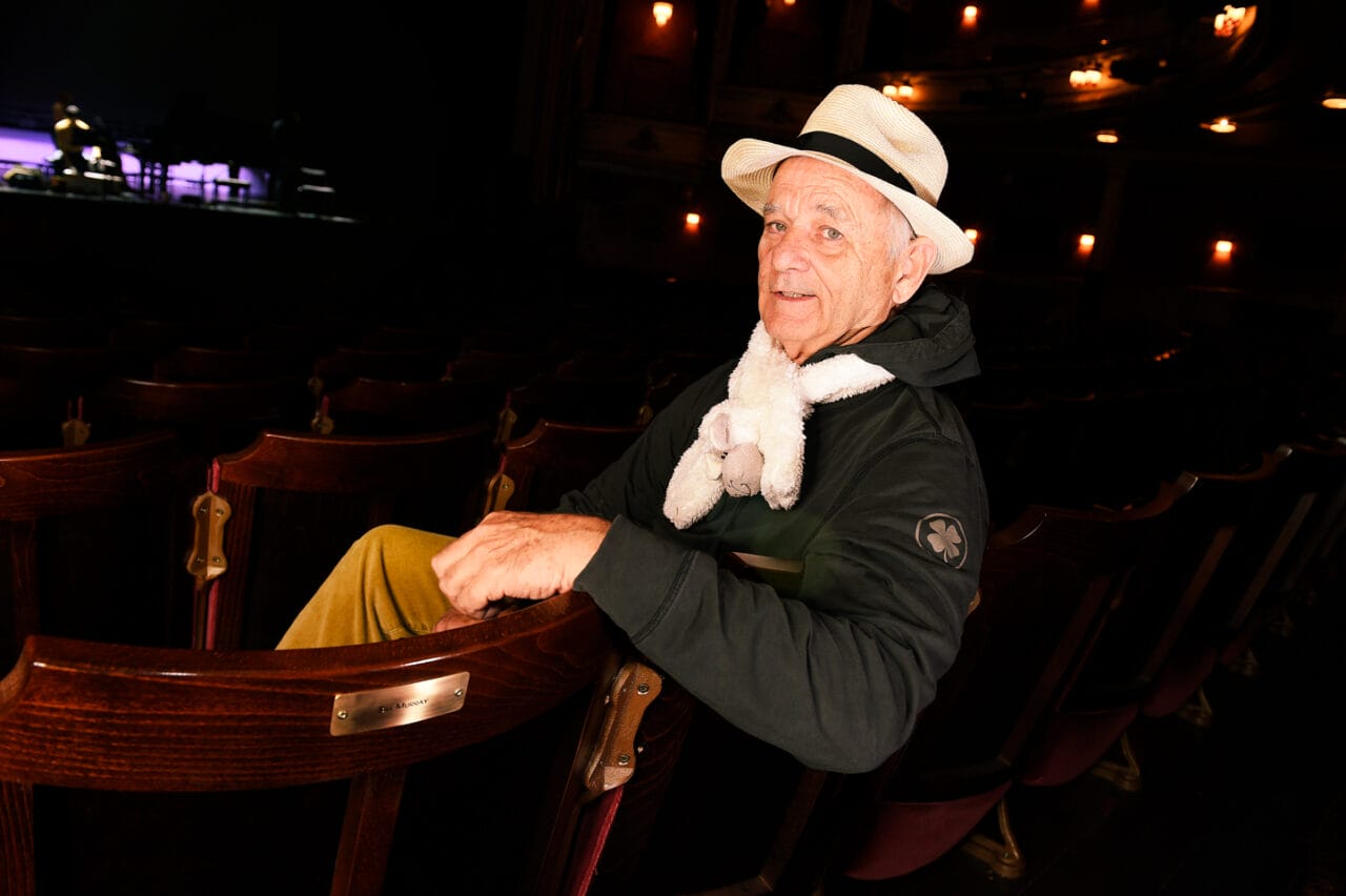 An older man wearing a hat and scarf sits in an empty theatre, looking back over his shoulder toward the camera.