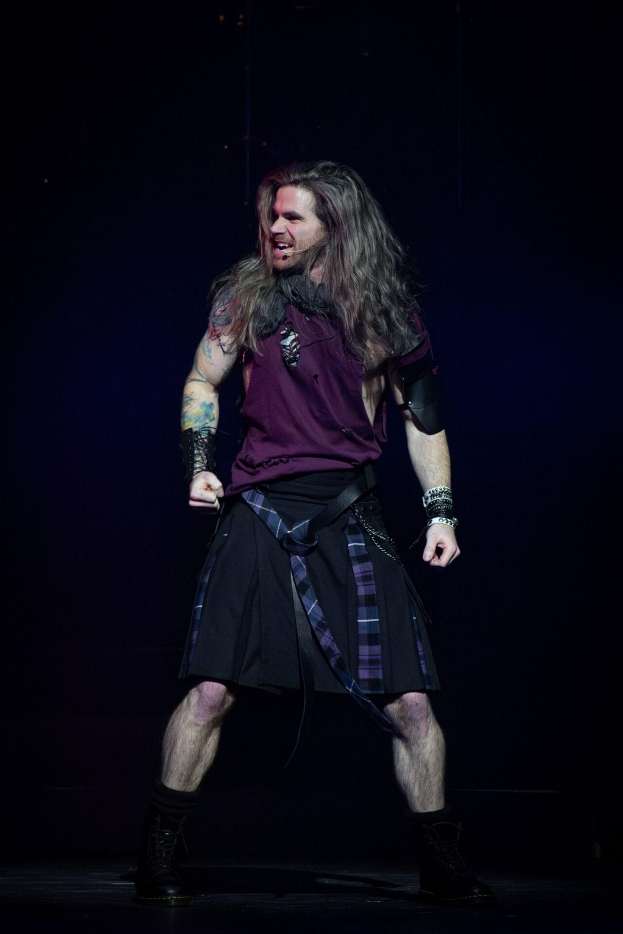 A person with long hair, wearing a sleeveless maroon shirt, a black kilt with a checked accent, and black boots, stands on stage with clenched fists and a fierce expression, ready to channel the energy of "We Will Rock You.