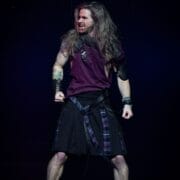 A person with long hair, wearing a sleeveless maroon shirt, a black kilt with a checked accent, and black boots, stands on stage with clenched fists and a fierce expression, ready to channel the energy of "We Will Rock You.