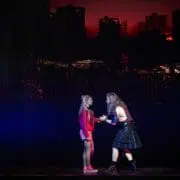 Two people stand on stage in dramatic lighting, the backdrop of damaged buildings against a red sky evoking a post-apocalyptic "We Will Rock You" vibe. One wears a red outfit and trainers; the other sports a kilt and boots.