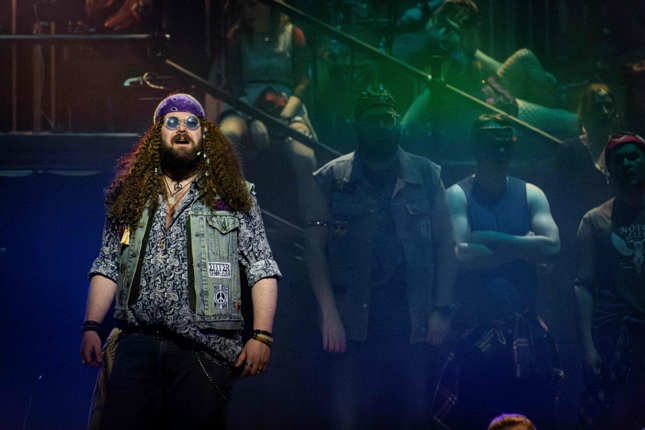A person with long curly hair, sunglasses, and a bandana stands on stage, channelling We Will Rock You vibes in a patterned shirt and waistcoat with patches. Several others are in the background under dramatic, coloured lighting.