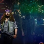 A person with long curly hair, sunglasses, and a bandana stands on stage, channelling We Will Rock You vibes in a patterned shirt and waistcoat with patches. Several others are in the background under dramatic, coloured lighting.