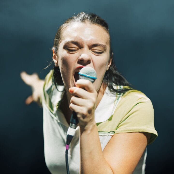 A person passionately singing into a microphone with eyes closed on stage, arm extended outward against a dark background.