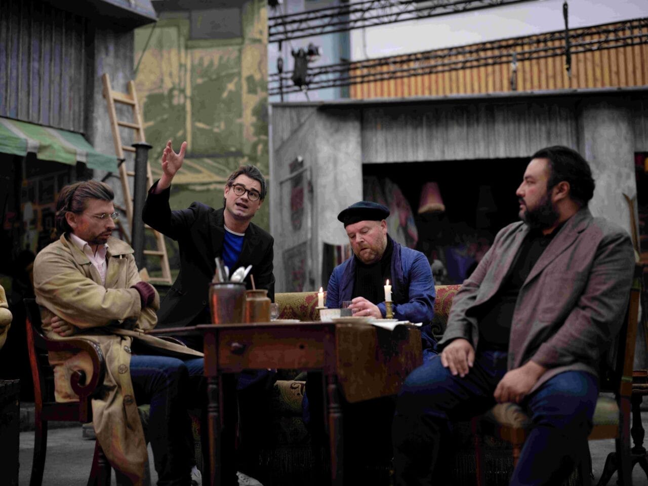 Four men sit around a table in an outdoor setting; one man gestures animatedly while the others listen or look away.