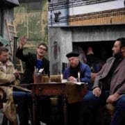 Four men sit around a table in an outdoor setting; one man gestures animatedly while the others listen or look away.
