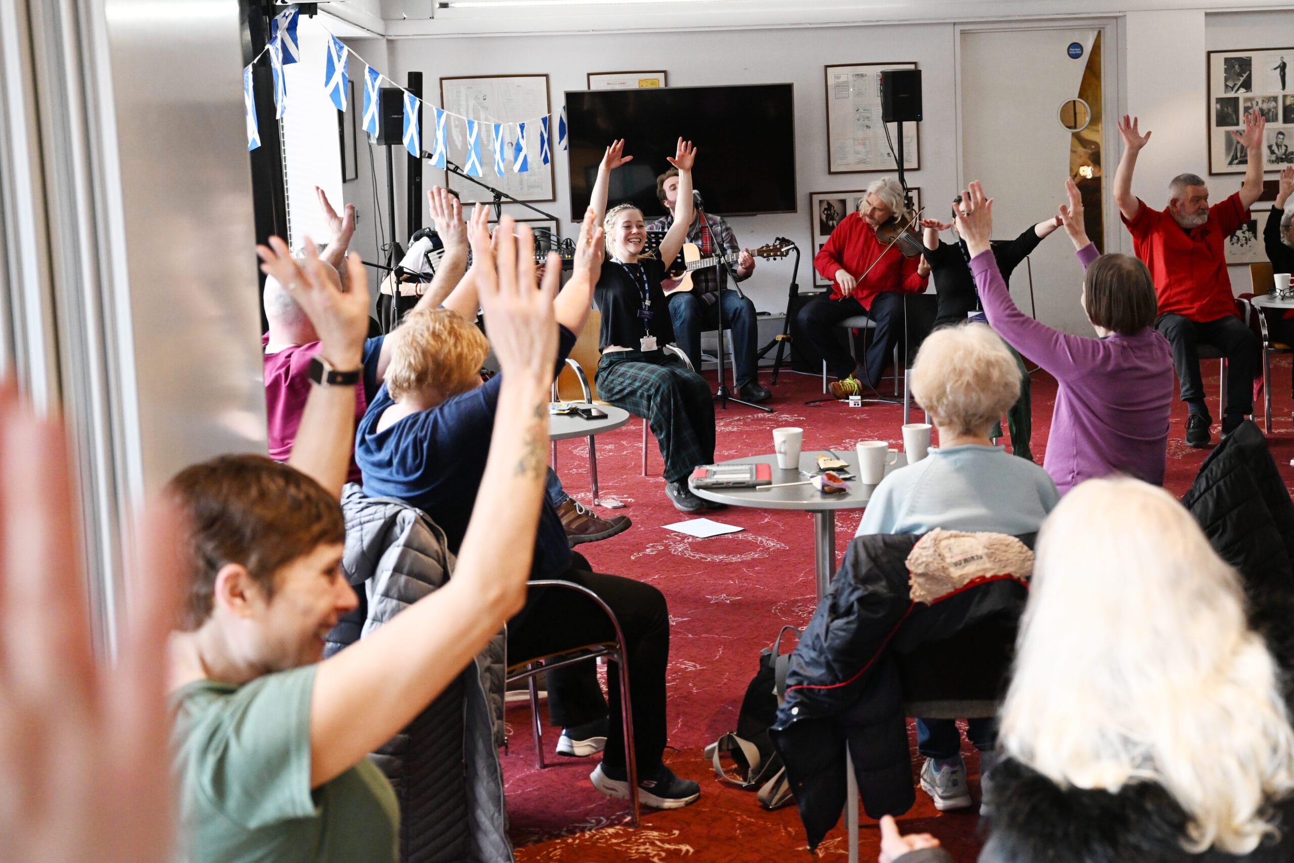 A group of older adults sit in a semicircle raising their arms during an indoor group activity; a few musicians are playing instruments at the front.