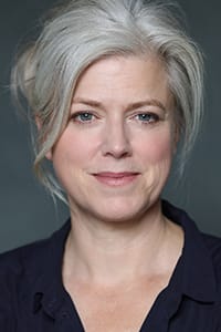 A woman with light gray hair in a loose updo, wearing a dark collared shirt, looks at the camera with a neutral expression against a dark background.