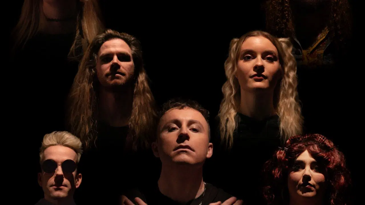 Seven people with serious expressions are positioned in a triangular arrangement against a black background, evoking the iconic "Bohemian Rhapsody" pose. Their faces are lit from below, casting dramatic shadows. The central person stands in front with arms folded.