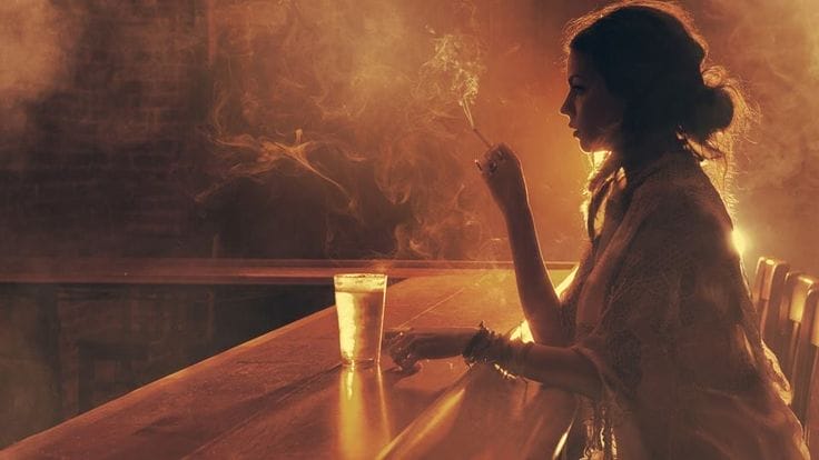 A woman sits at a bar holding a cigarette with a glass in front of her, illuminated by warm, soft lighting and surrounded by smoke.