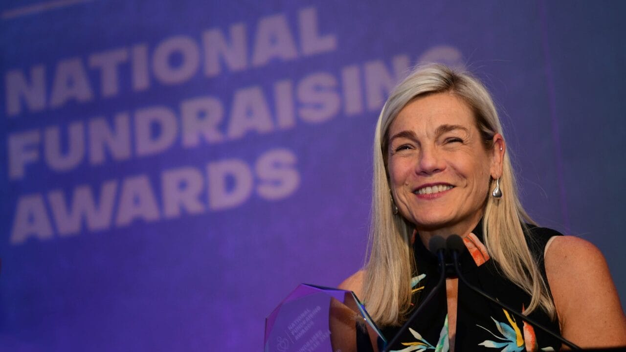 A woman stands at a podium smiling and holding an award, with a "National Fundraising Awards" sign in the background.