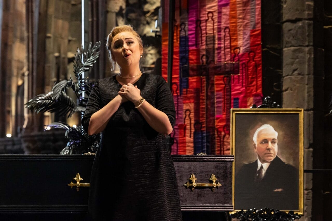 A woman in a black dress sings in front of a closed black casket and a portrait of an older man, evoking the dramatic flair of L'heure espagnole, with a decorative backdrop in a dimly lit room.