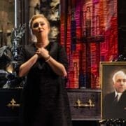 A woman in a black dress sings in front of a closed black casket and a portrait of an older man, evoking the dramatic flair of L'heure espagnole, with a decorative backdrop in a dimly lit room.