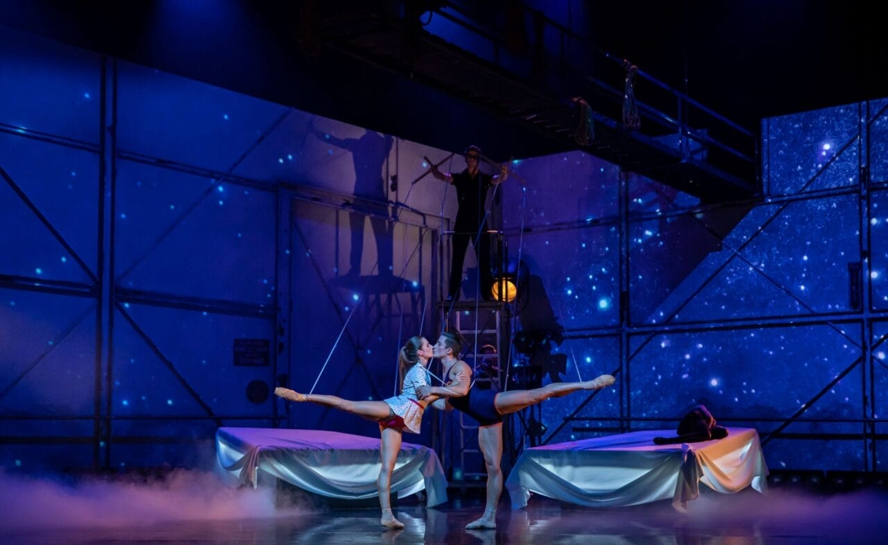 Two ballet dancers perform an acrobatic lift on stage, with blue lighting, starry backdrop, and another performer on a platform above them.