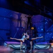 Two ballet dancers perform an acrobatic lift on stage, with blue lighting, starry backdrop, and another performer on a platform above them.