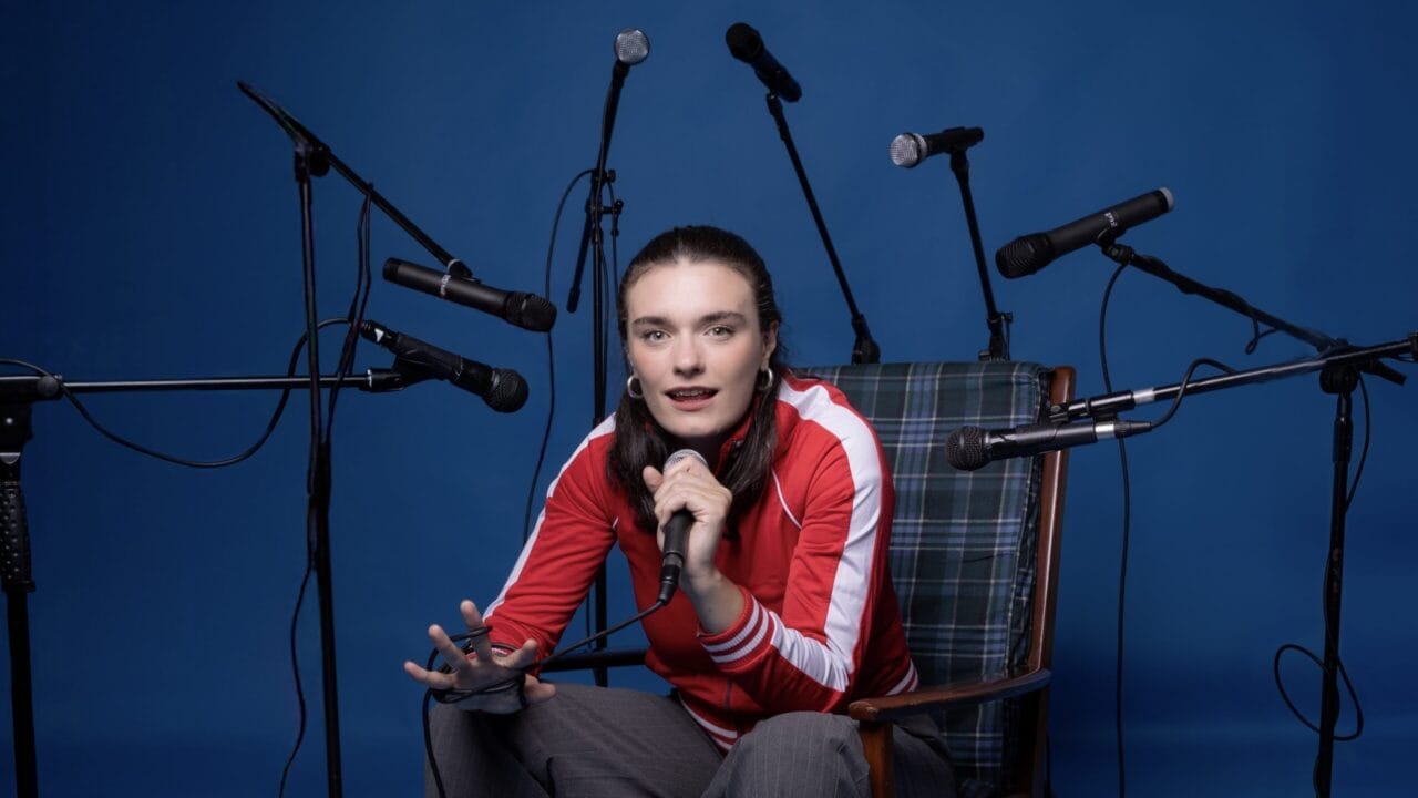 A person in a red jacket sits on a chair, holding a microphone, surrounded by several other microphones against a blue background—ready to share their story in the Common Tongue.