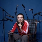 A person in a red jacket sits on a chair, holding a microphone, surrounded by several other microphones against a blue background—ready to share their story in the Common Tongue.