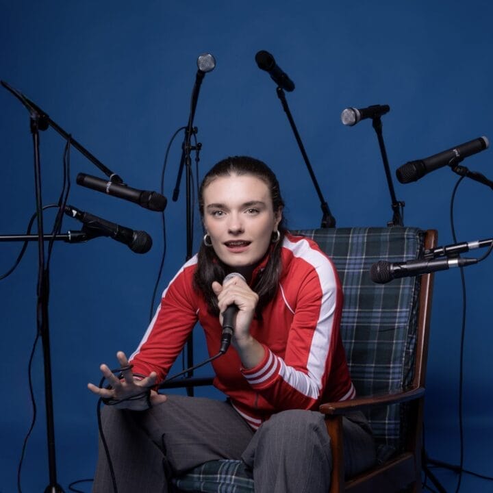 A person in a red jacket sits on a chair, holding a microphone, surrounded by several other microphones against a blue background—ready to share their story in the Common Tongue.