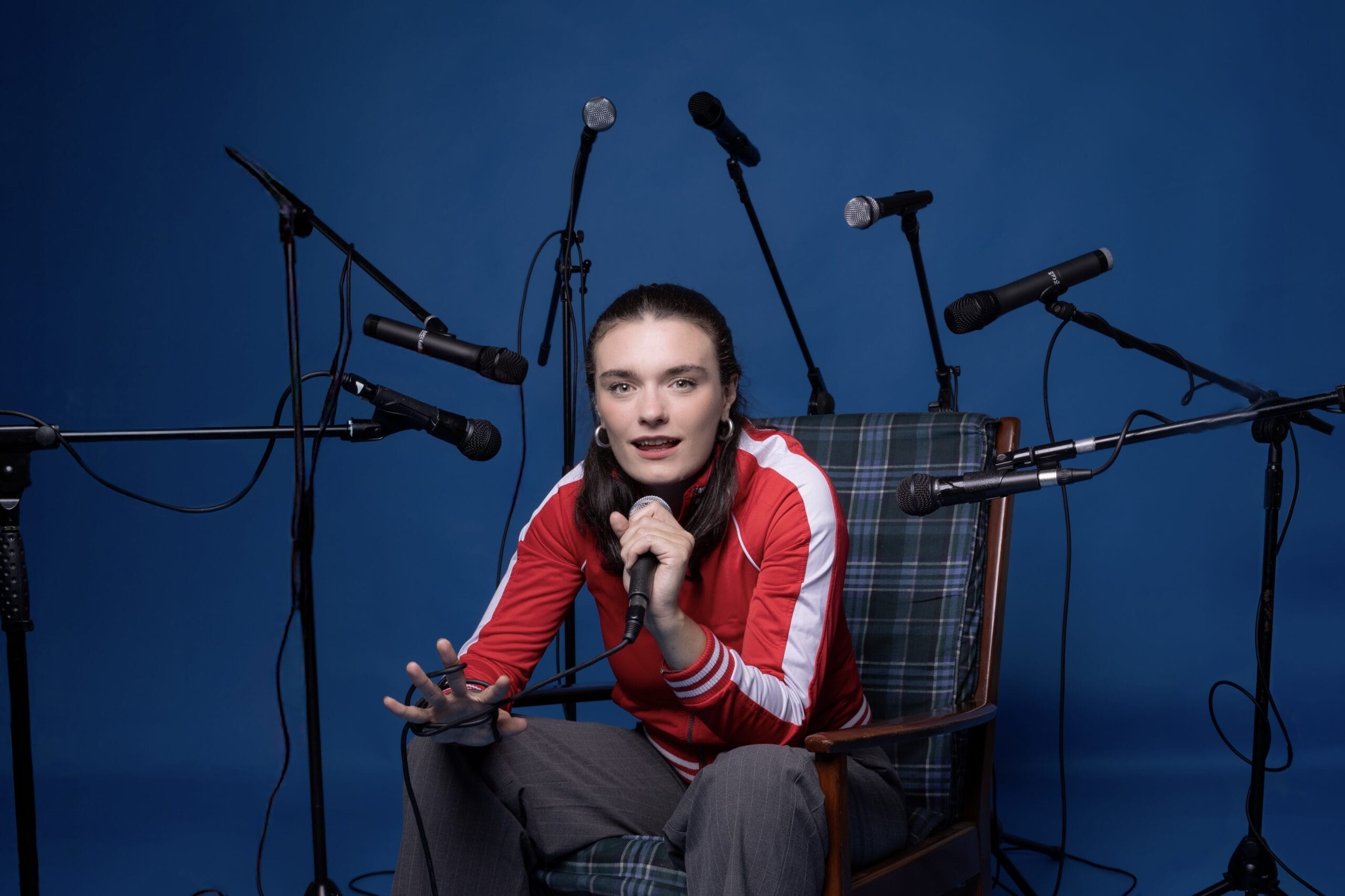 A person in a red jacket sits on a chair, holding a microphone, surrounded by several other microphones against a blue background—ready to share their story in the Common Tongue.