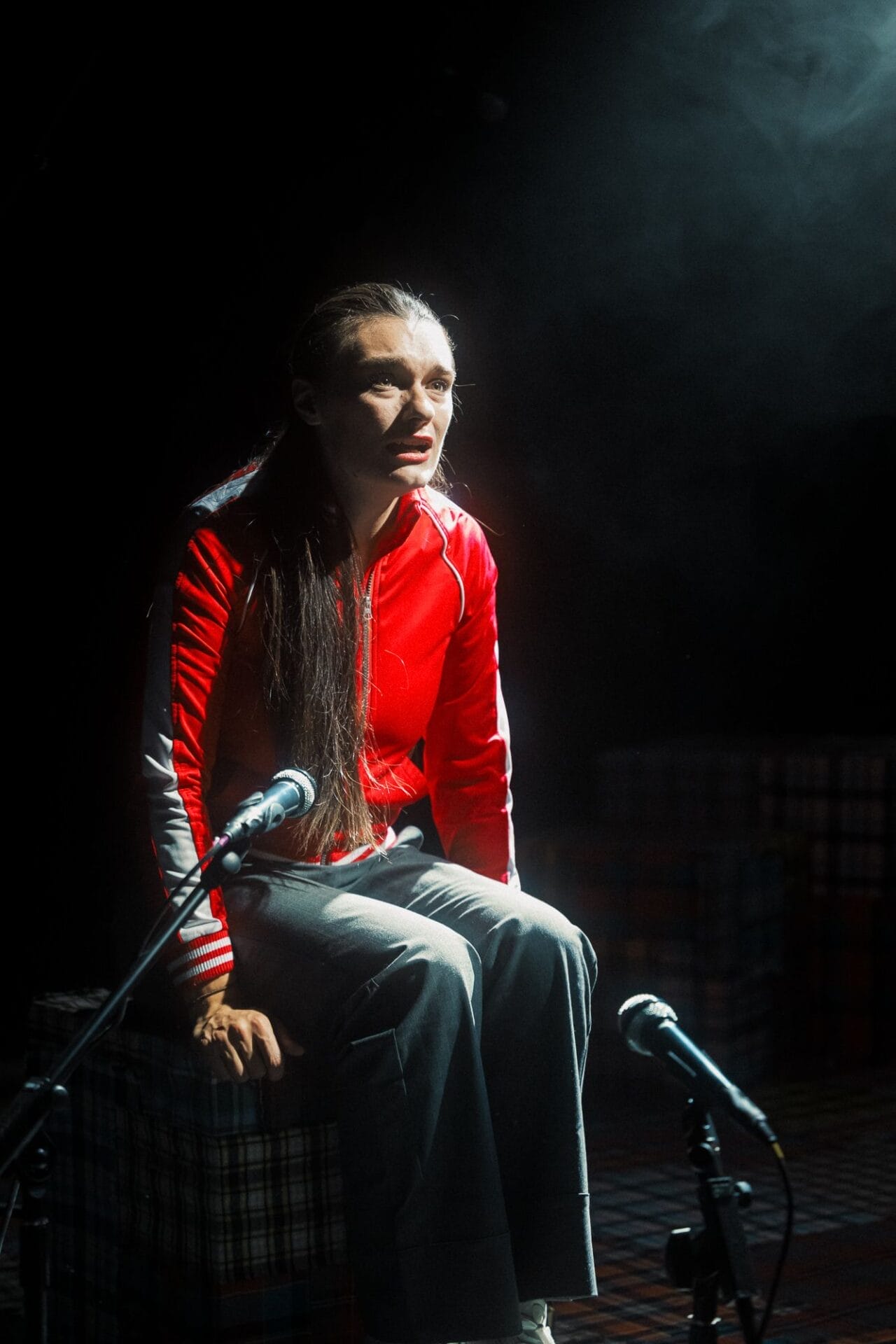A woman in a red jacket and gray pants sits on a stage, lit by a spotlight, with two microphones in front of her and a smoky background, poised to share her story in the Common Tongue.