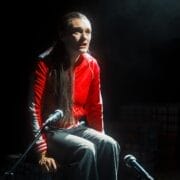 A woman in a red jacket and gray pants sits on a stage, lit by a spotlight, with two microphones in front of her and a smoky background, poised to share her story in the Common Tongue.